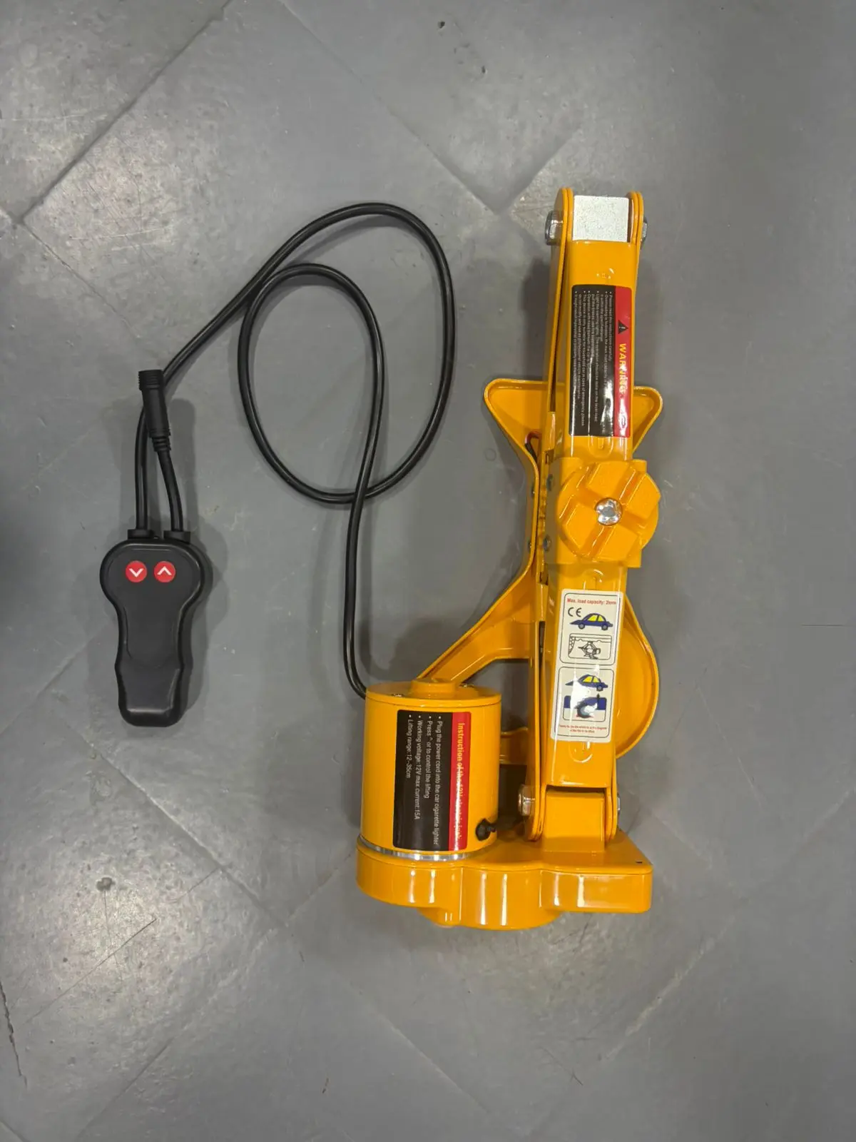 Gato hidráulico eléctrico 12V automático color amarillo con control remoto negro de dos botones, sobre piso de baldosa gris.