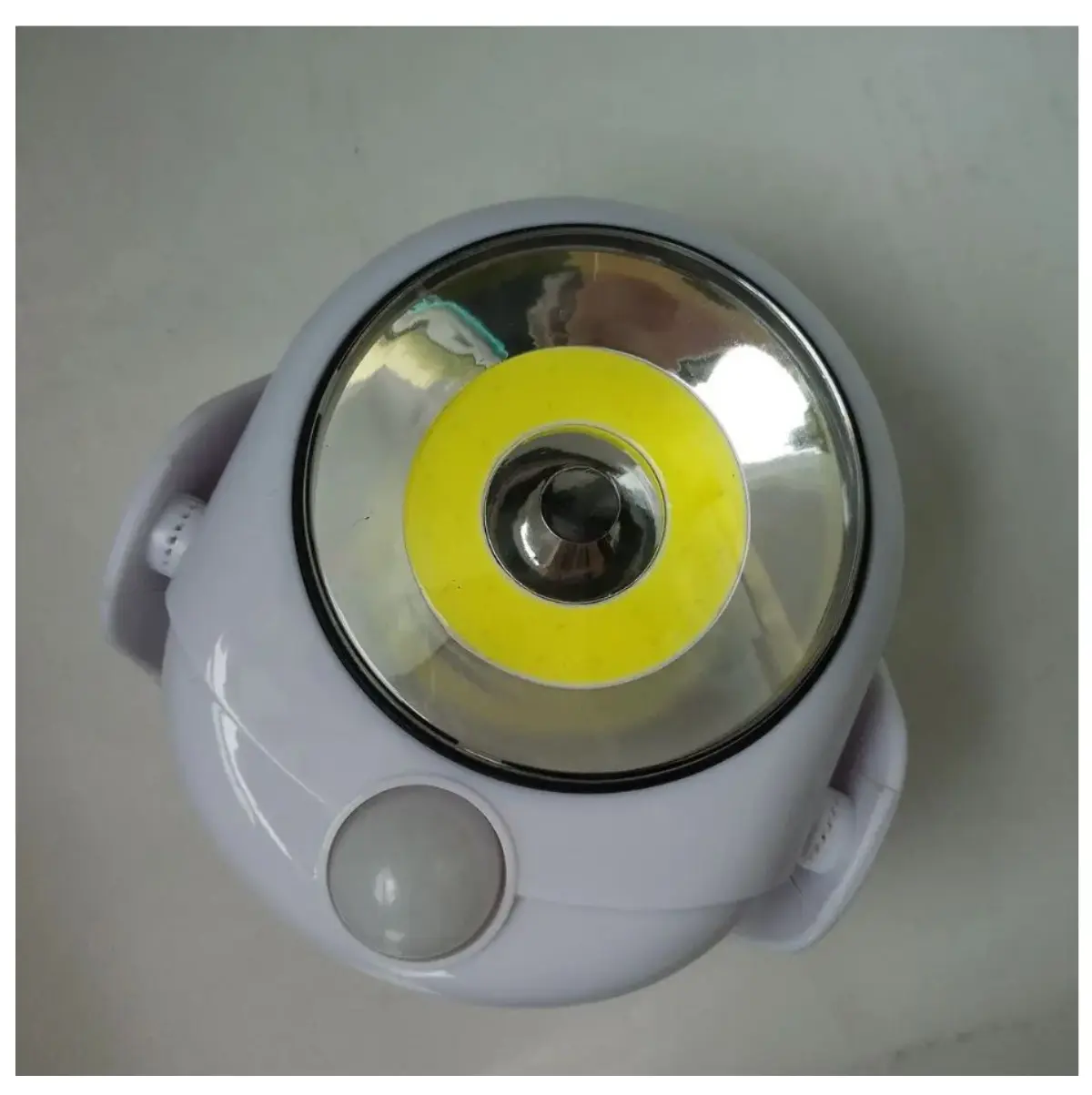 Lámpara LED blanca esférica con lente de anillo amarillo, sensor de movimiento y soportes laterales, vista desde arriba.