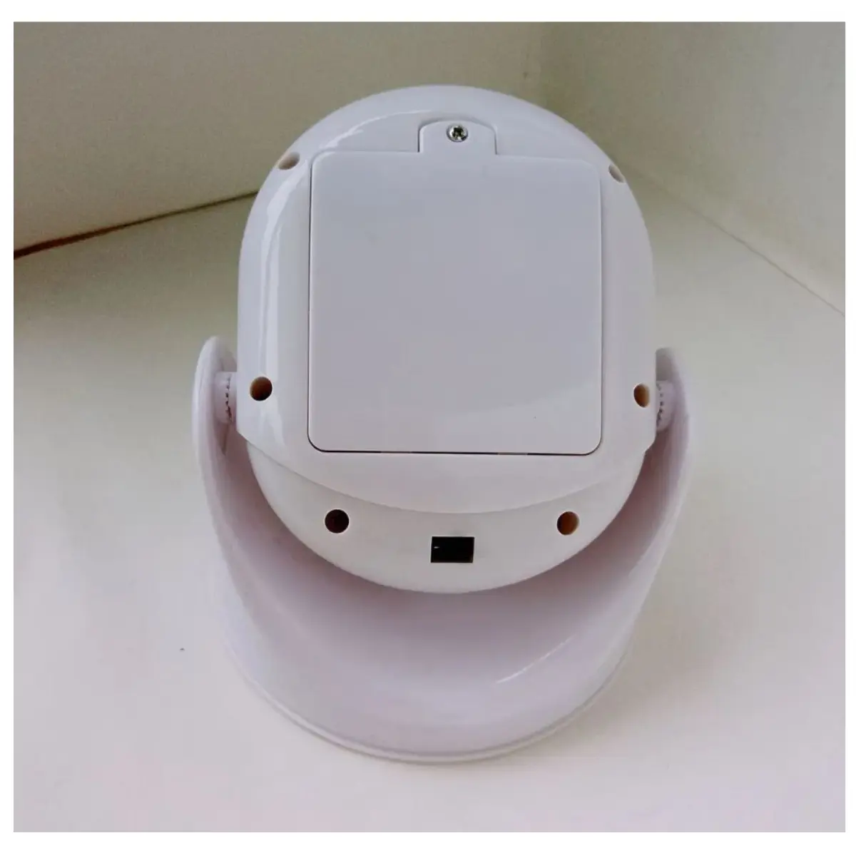 Vista posterior de lámpara LED blanca con sensor, mostrando compartimento de baterías, interruptor de encendido y base giratoria sobre superficie plana.