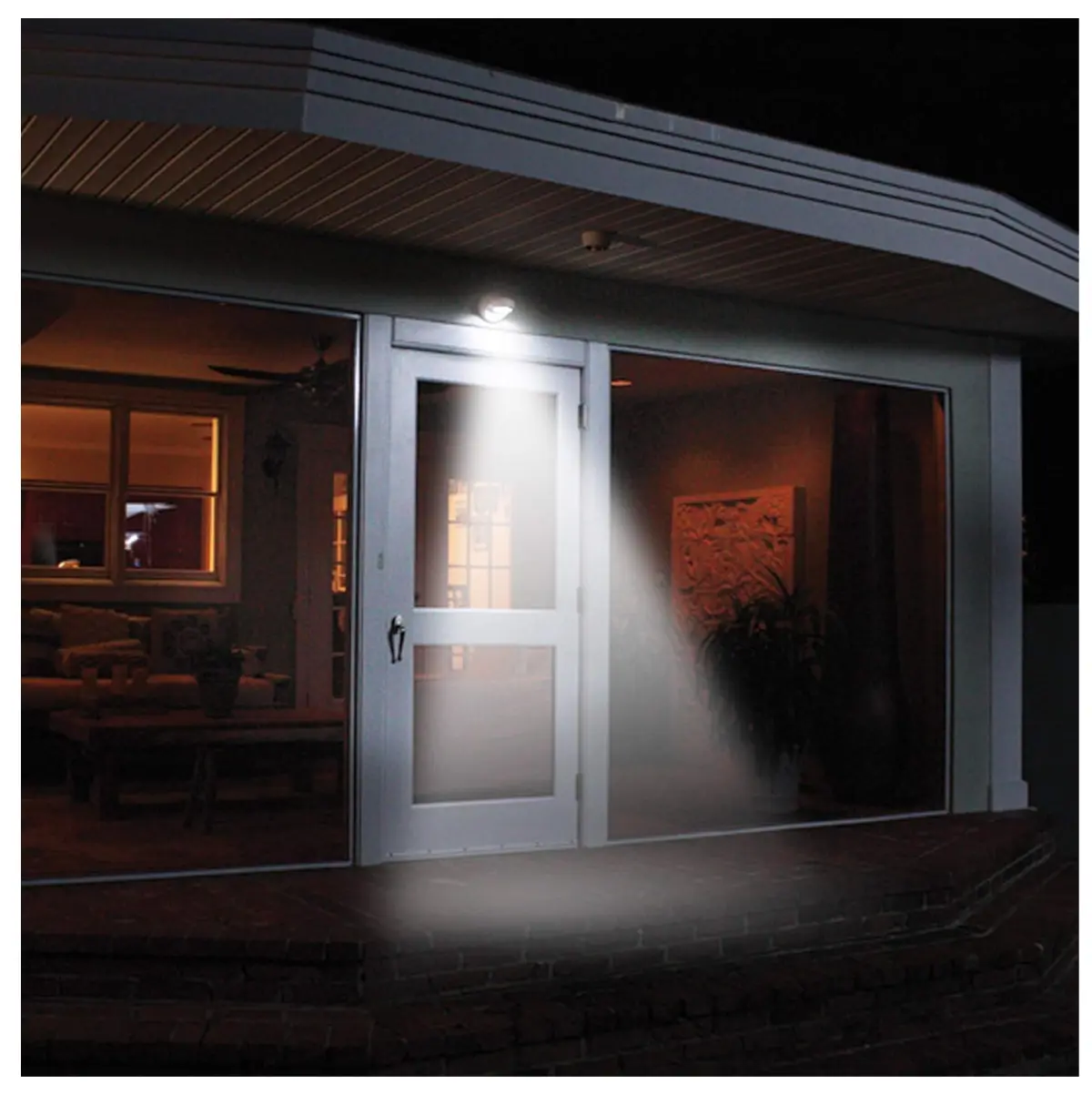 Lámpara LED blanca con sensor de movimiento instalada sobre una puerta de entrada, emitiendo un haz de luz brillante hacia el suelo en un entorno nocturno.