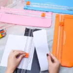 Guillotinas para papel con cuchilla oculta en colores azul, rosa y naranja, junto a una base negra en uso para cortar hojas blancas sobre mesa de mármol.
