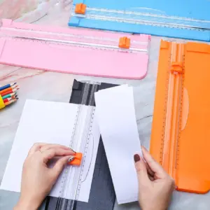 Guillotinas para papel con cuchilla oculta en colores azul, rosa y naranja, junto a una base negra en uso para cortar hojas blancas sobre mesa de mármol.