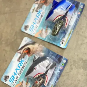 Dos tiburones de juguete en su empaque: uno azul y otro gris, ambos con carro de metal atrapado en sus fauces mecánicas.