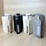 Cuatro termos portátiles para café de 350 ml con correa de tela, en colores crema, negro, blanco y gris, sobre una superficie de madera.
