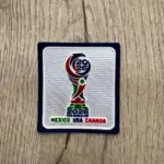Parche termoadhesivo del Mundial 2026 sobre madera, con trofeo bordado en rojo, verde y azul, y texto inferior que dice México USA Canadá.
