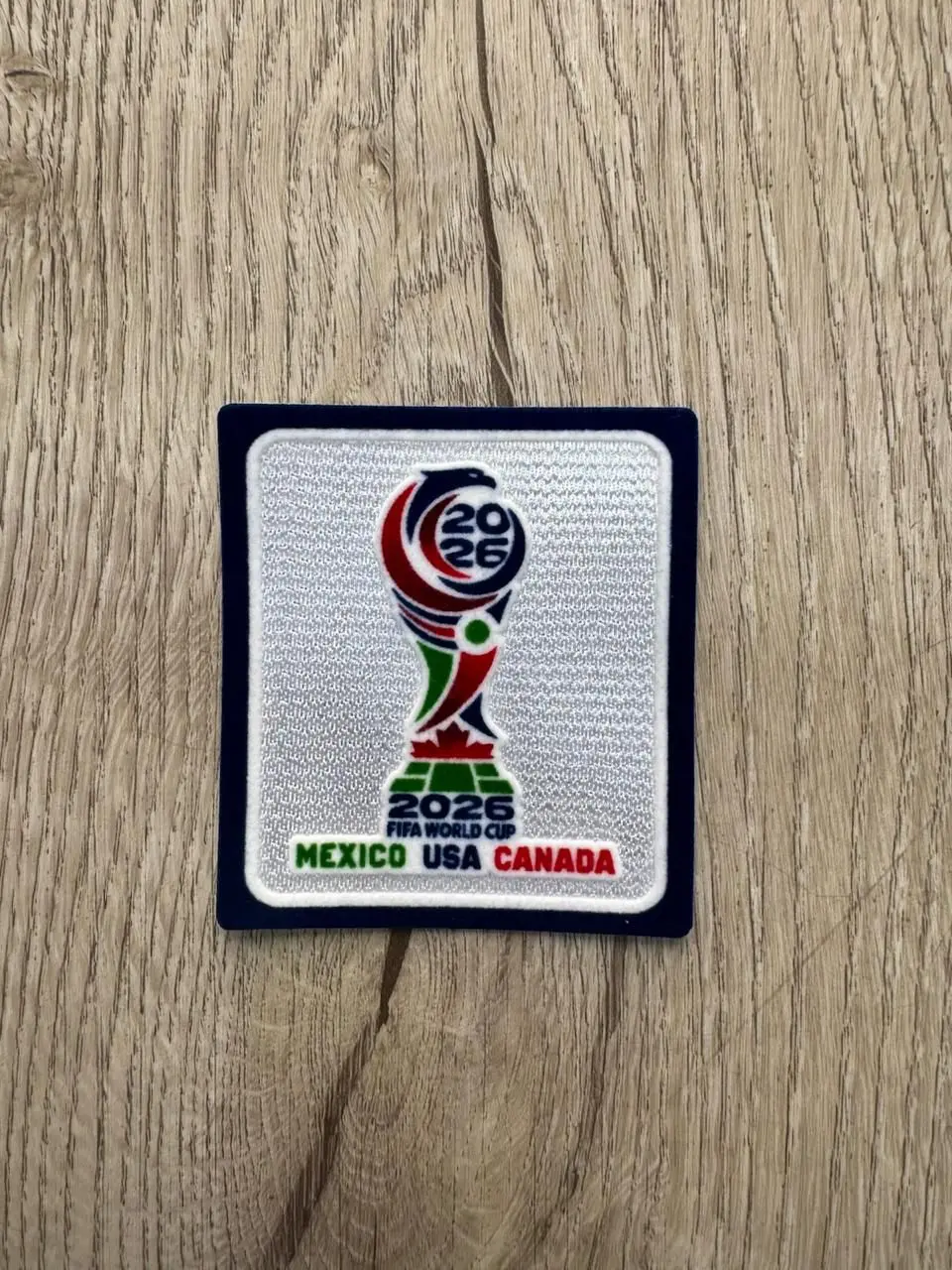 Parche termoadhesivo del Mundial 2026 sobre madera, con trofeo bordado en rojo, verde y azul, y texto inferior que dice México USA Canadá.
