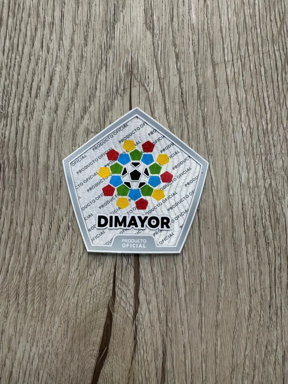 Parche termoadhesivo oficial DIMAYOR pentagonal sobre superficie de madera, con diseño colorido de balón y polígonos alrededor.