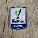 Parche termoadhesivo oficial de la Liga BetPlay Dimayor sobre superficie de madera, con diseño en azul, blanco, verde y amarillo.