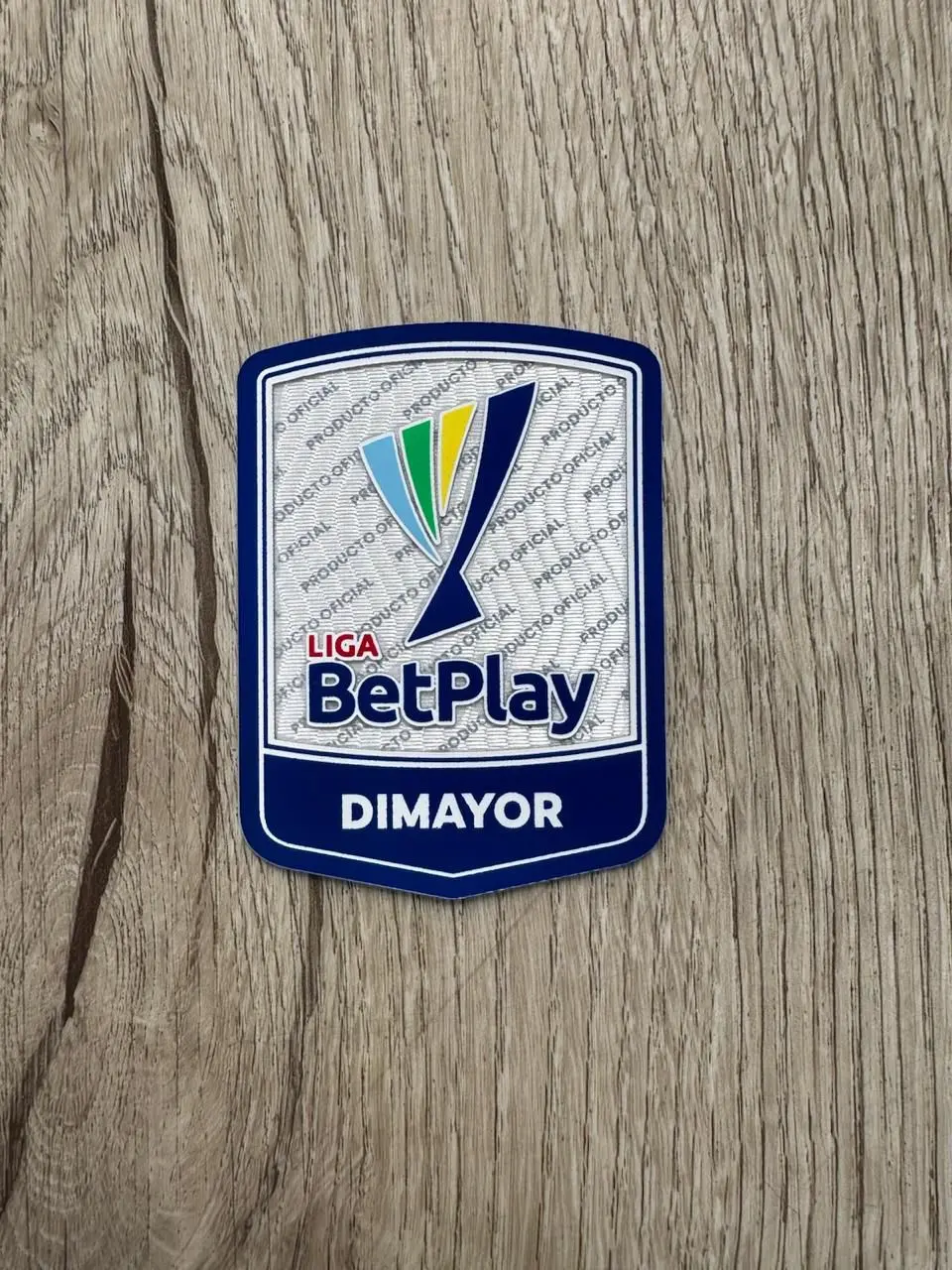Parche termoadhesivo oficial de la Liga BetPlay Dimayor sobre superficie de madera, con diseño en azul, blanco, verde y amarillo.