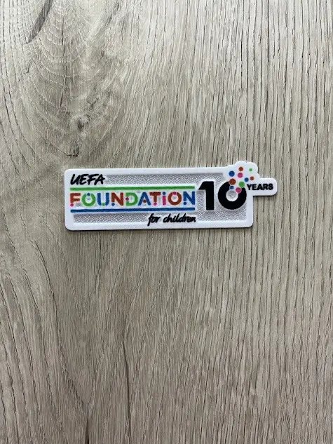 Parche termoadhesivo blanco rectangular de UEFA Foundation for Children 10 Years con letras coloridas, sobre superficie de madera.