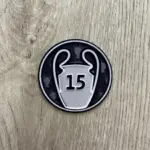 Parche termoadhesivo redondo con diseño plateado y negro del trofeo de la Champions League y el número 15, sobre superficie de madera.