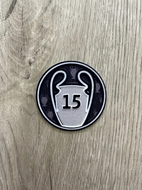 Parche termoadhesivo redondo con diseño plateado y negro del trofeo de la Champions League y el número 15, sobre superficie de madera.