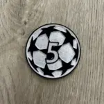 Parche termoadhesivo redondo con el número 5 blanco sobre diseño de balón de fútbol en blanco y negro, bordado sobre fondo de madera.