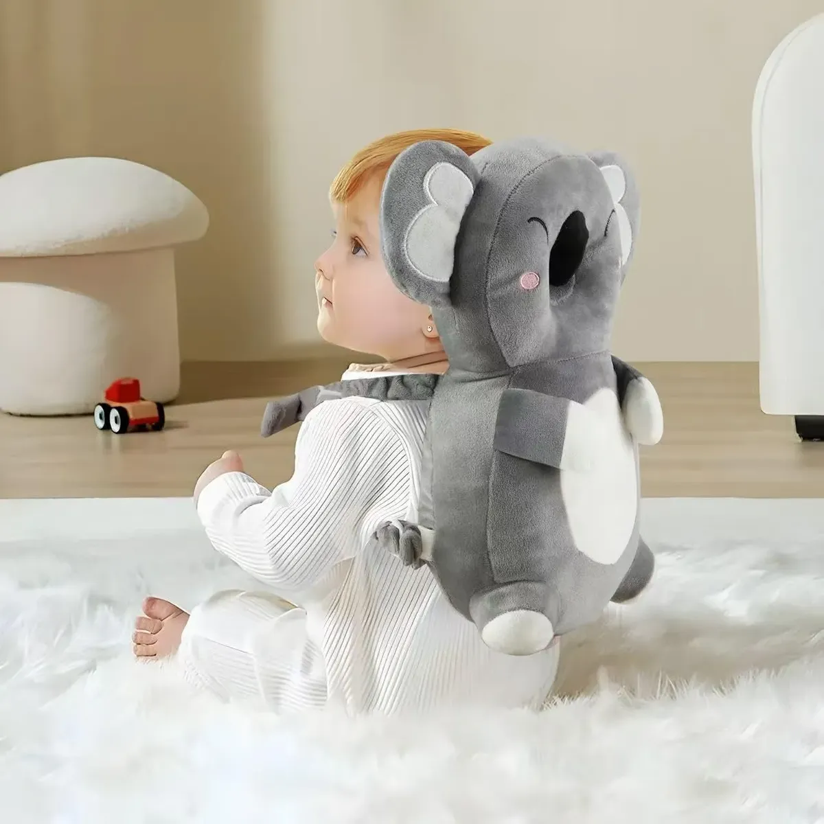 Almohada anticaída de peluche gris con diseño de koala, usada en la espalda de un bebé sentado sobre una alfombra blanca suave.