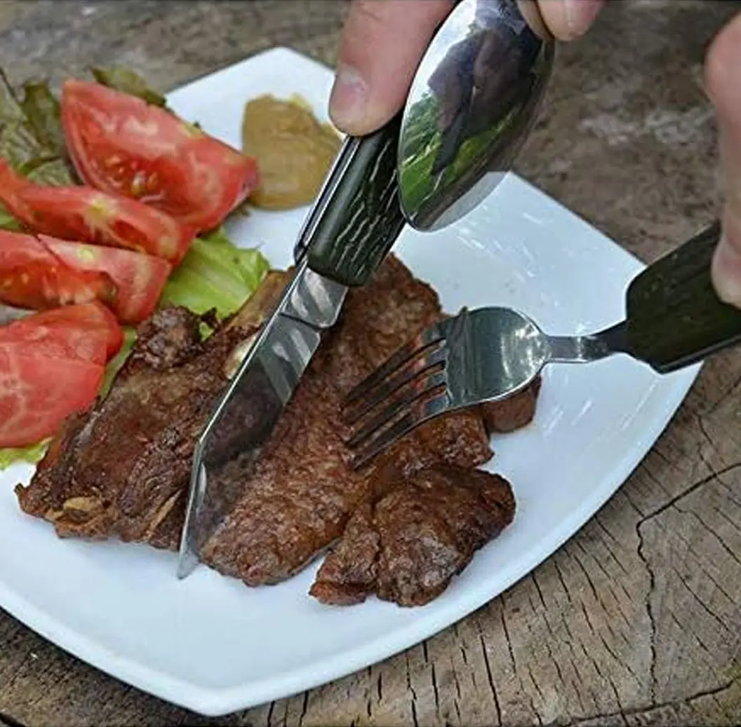 Set de cubiertos de camping plegables de acero inoxidable con mango negro, cortando carne en un plato con ensalada sobre mesa de madera.