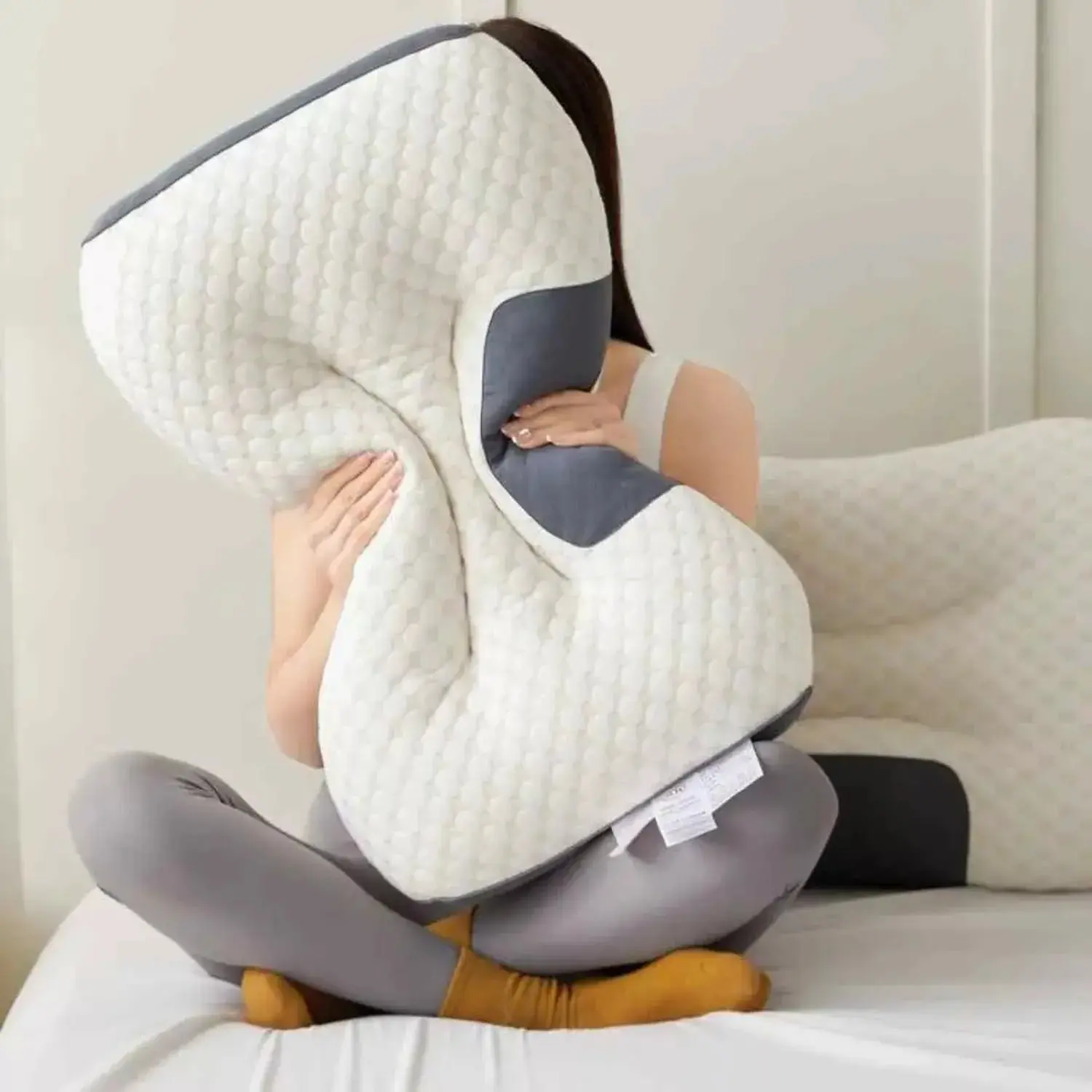 Almohada ergonómica 2x1 blanca con textura acolchada y detalles grises, abrazada por una mujer en cama para mostrar su soporte corporal.