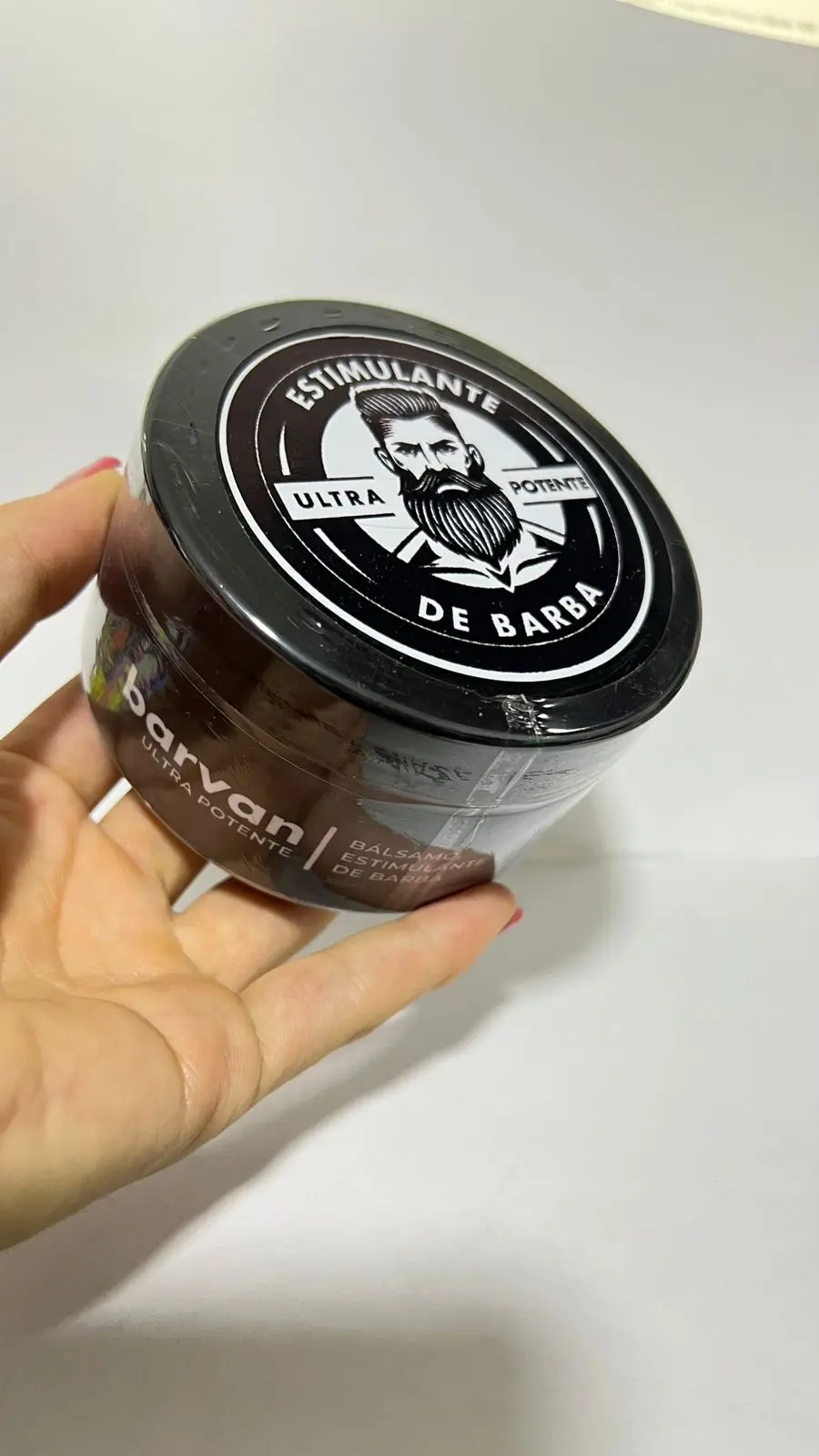 Mano sosteniendo tarro redondo negro de bálsamo estimulante para barba con etiqueta circular y diseño de hombre barbudo
