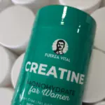 Envase verde cilíndrico de creatina monohidratada para mujer con logotipo de hombre con bigote, rodeado de tarros blancos. Contenido 240g.