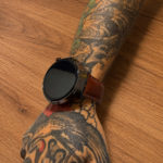 Reloj inteligente Vortexx Smart-Active con pantalla negra y correa de cuero marrón, usado en un brazo con tatuajes sobre fondo de madera.