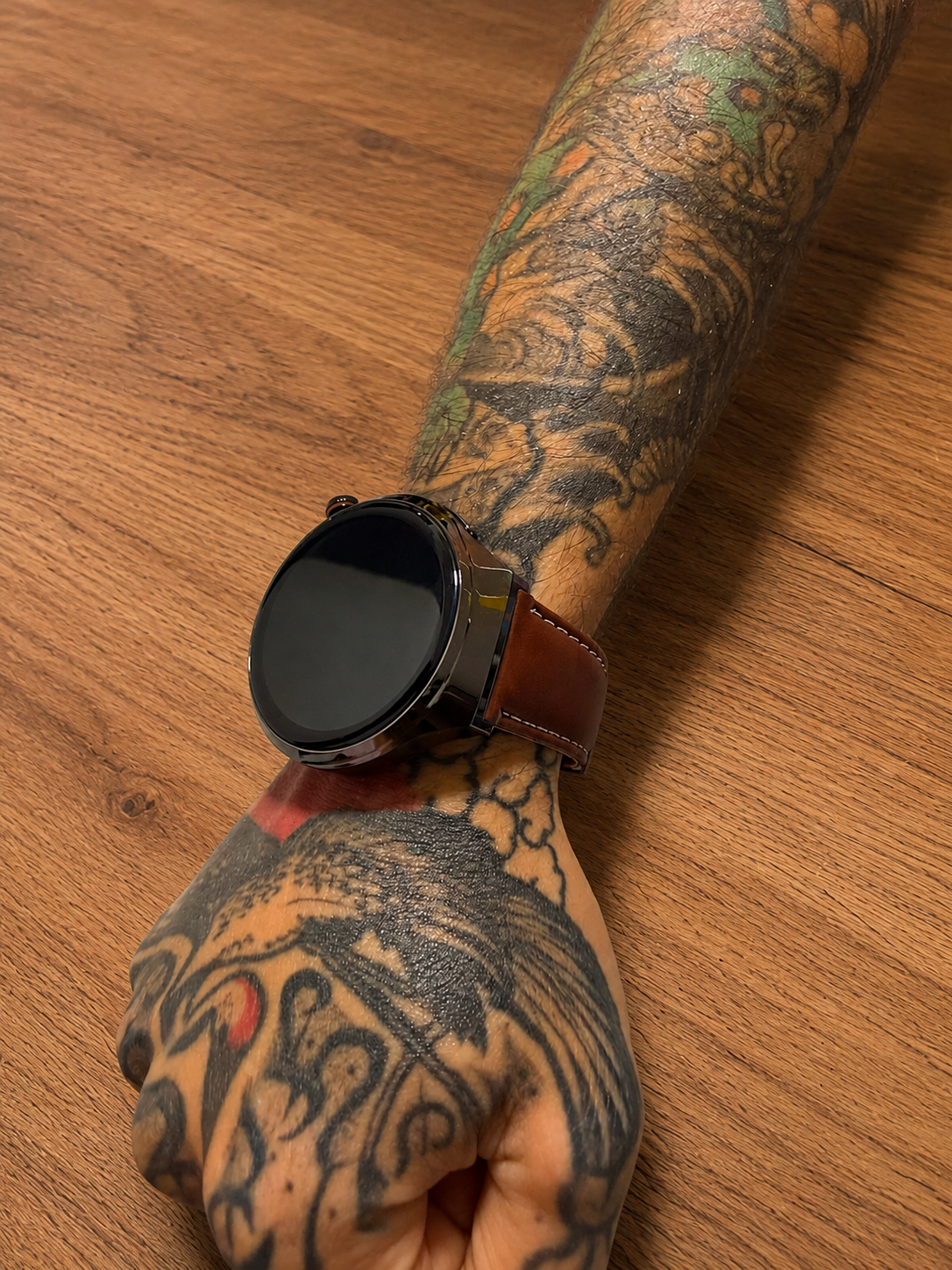 Reloj inteligente Vortexx Smart-Active con pantalla negra y correa de cuero marrón, usado en un brazo con tatuajes sobre fondo de madera.