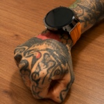 Reloj inteligente Vortexx Smart-Active negro con esfera redonda y correa naranja de silicona, colocado sobre una muñeca tatuada.