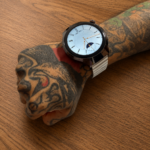 Reloj inteligente Vortexx Smart-Active con caja negra, esfera analógica y correa metálica blanca en una muñeca tatuada sobre madera.
