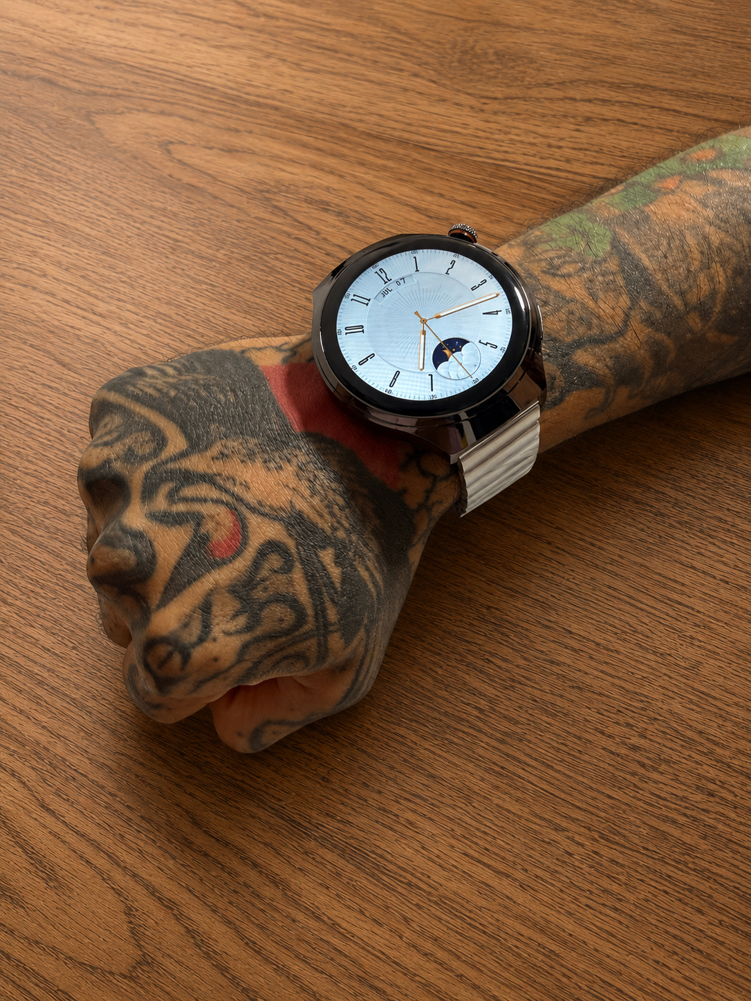 Reloj inteligente Vortexx Smart-Active con caja negra, esfera analógica y correa metálica blanca en una muñeca tatuada sobre madera.