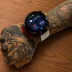 Reloj inteligente Vortexx Smart-Active negro con pantalla redonda y correa metálica flexible, usado en muñeca tatuada sobre mesa de madera.