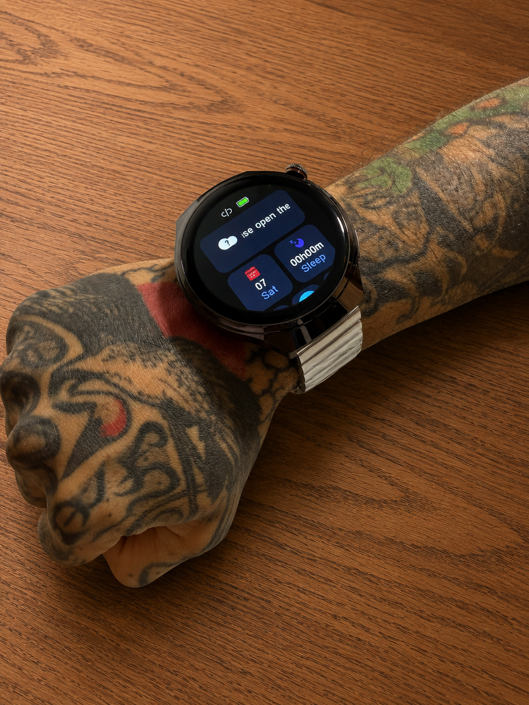 Reloj inteligente Vortexx Smart-Active negro con pantalla redonda y correa metálica flexible, usado en muñeca tatuada sobre mesa de madera.