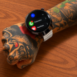 Reloj inteligente con esfera circular negra y correa metálica plateada, colocado sobre un brazo tatuado con un menú digital visible en la pantalla.