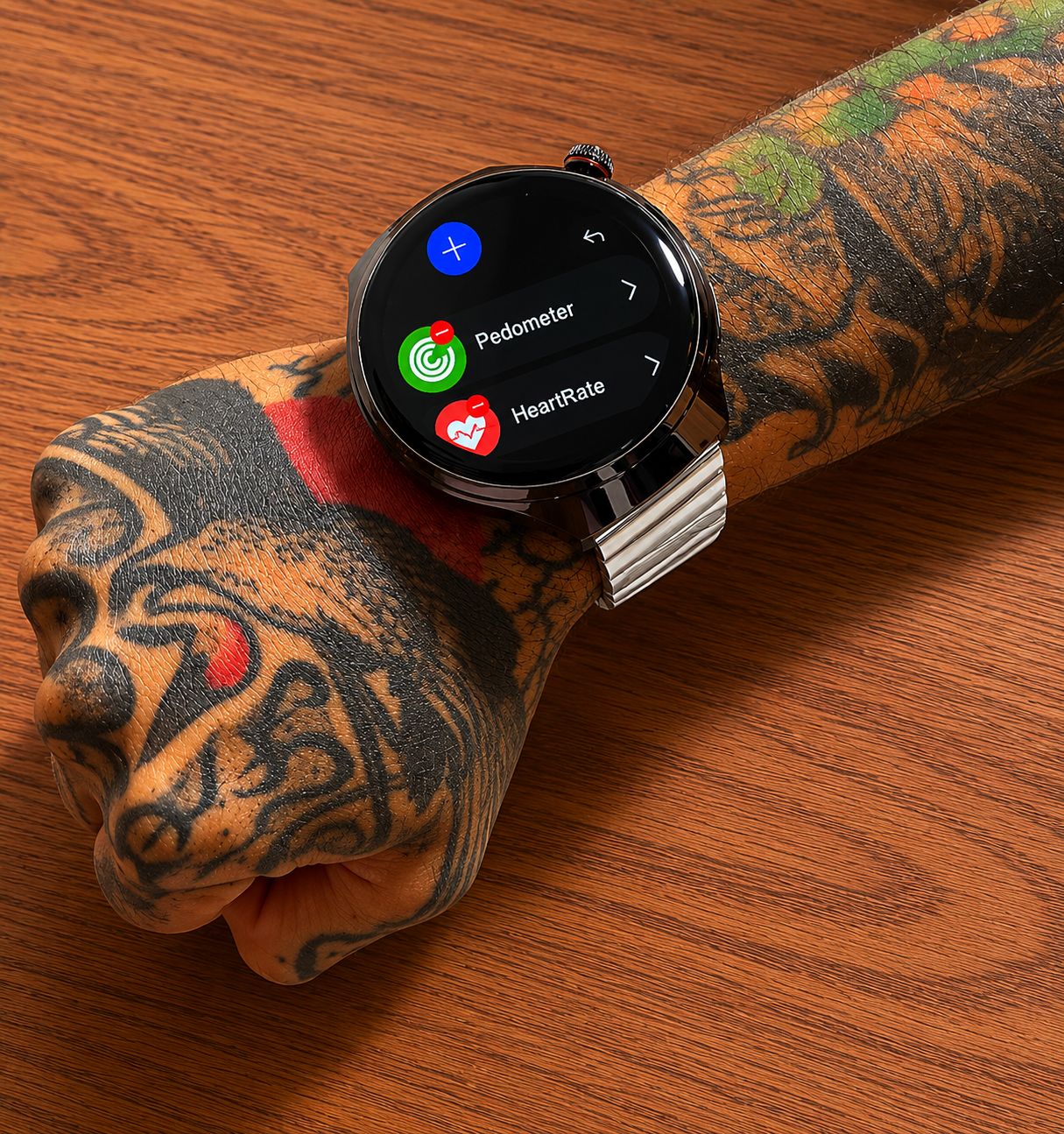 Reloj inteligente con esfera circular negra y correa metálica plateada, colocado sobre un brazo tatuado con un menú digital visible en la pantalla.