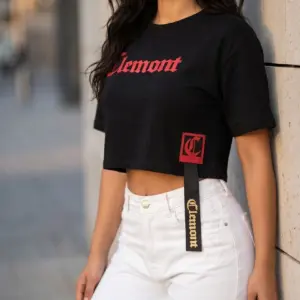 Blusa crop oversize negra de algodón con texto rojo "Clemont" en el pecho, parche cuadrado con la letra C y cinta colgante.
