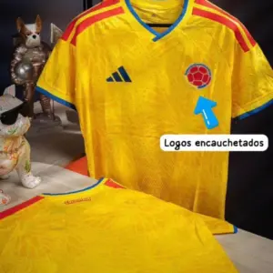 Camiseta de fútbol Colombia amarilla, versión jugador con logos encauchetados, detalles rojos y azules, mostrada colgada y doblada.