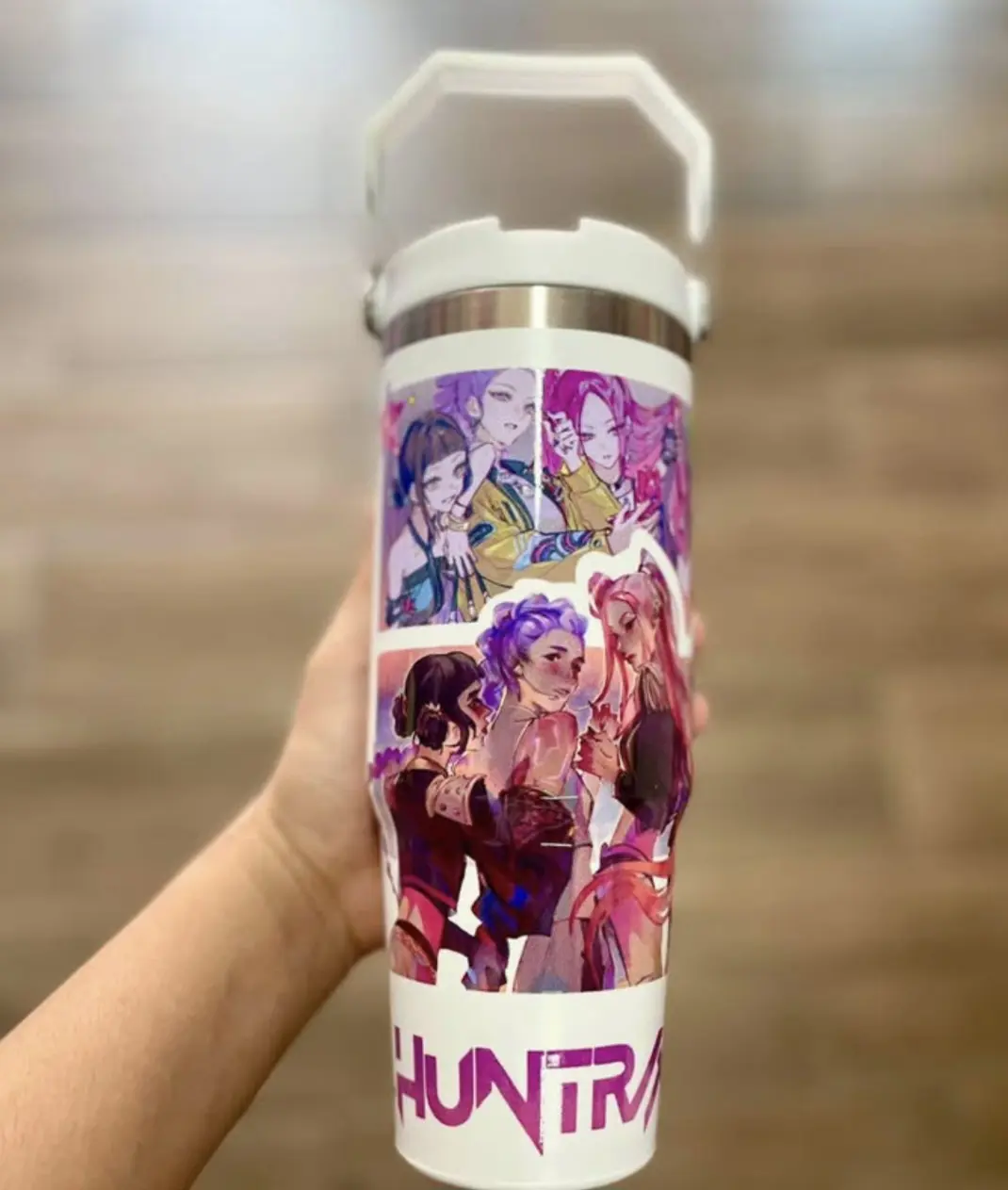 Termo de acero inoxidable blanco con asa, decorado con pegatinas de estilo anime K-pop y el texto HUNTR en la base, sostenido por una mano.