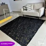 Alfombra negra con patrón de líneas doradas metalizadas, dispuesta en una sala con piso claro y sofá beige, aportando elegancia al ambiente.