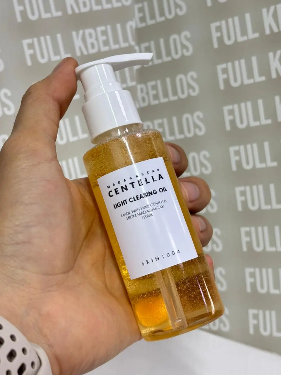 Mano sosteniendo botella transparente de 120ml con aceite limpiador facial amarillo, dispensador blanco y etiqueta blanca con letras negras.