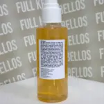 Envase transparente con bomba dispensadora blanca que contiene aceite limpiador facial dorado y etiqueta posterior con ingredientes en fondo blanco.
