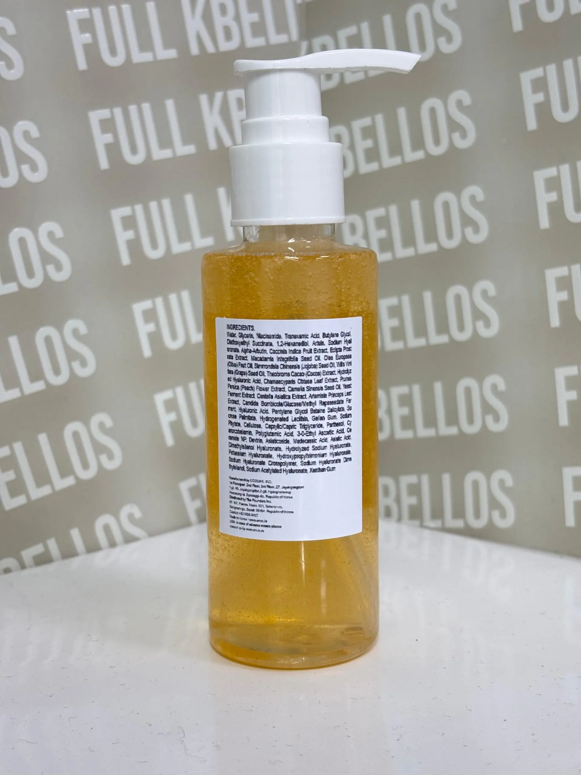 Envase transparente con bomba dispensadora blanca que contiene aceite limpiador facial dorado y etiqueta posterior con ingredientes en fondo blanco.