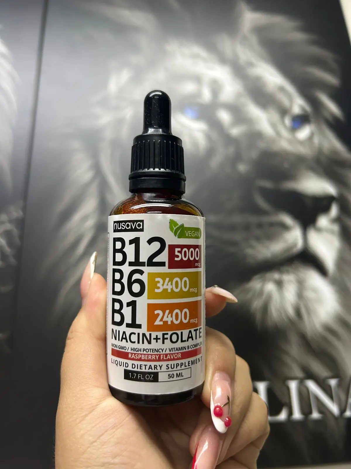 Mano sosteniendo gotero con complejo B líquido vegano de 50 ml y sabor a frambuesa frente a un póster en blanco y negro.