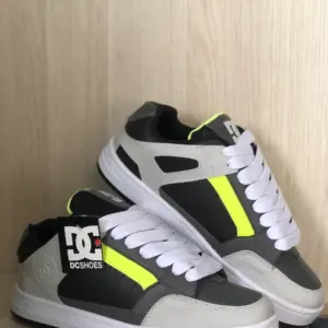 Tenis DC Shoes de skate en gris, negro y blanco con detalles amarillo neón, cordones blancos y suela gruesa sobre fondo de madera clara.