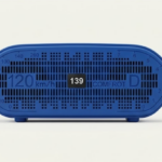 Parlante mini inalámbrico de color azul brillante, diseño rectangular con rejilla perforada, pantalla digital central con número 139, escalas de velocidad impresas en los bordes y patas de soporte.