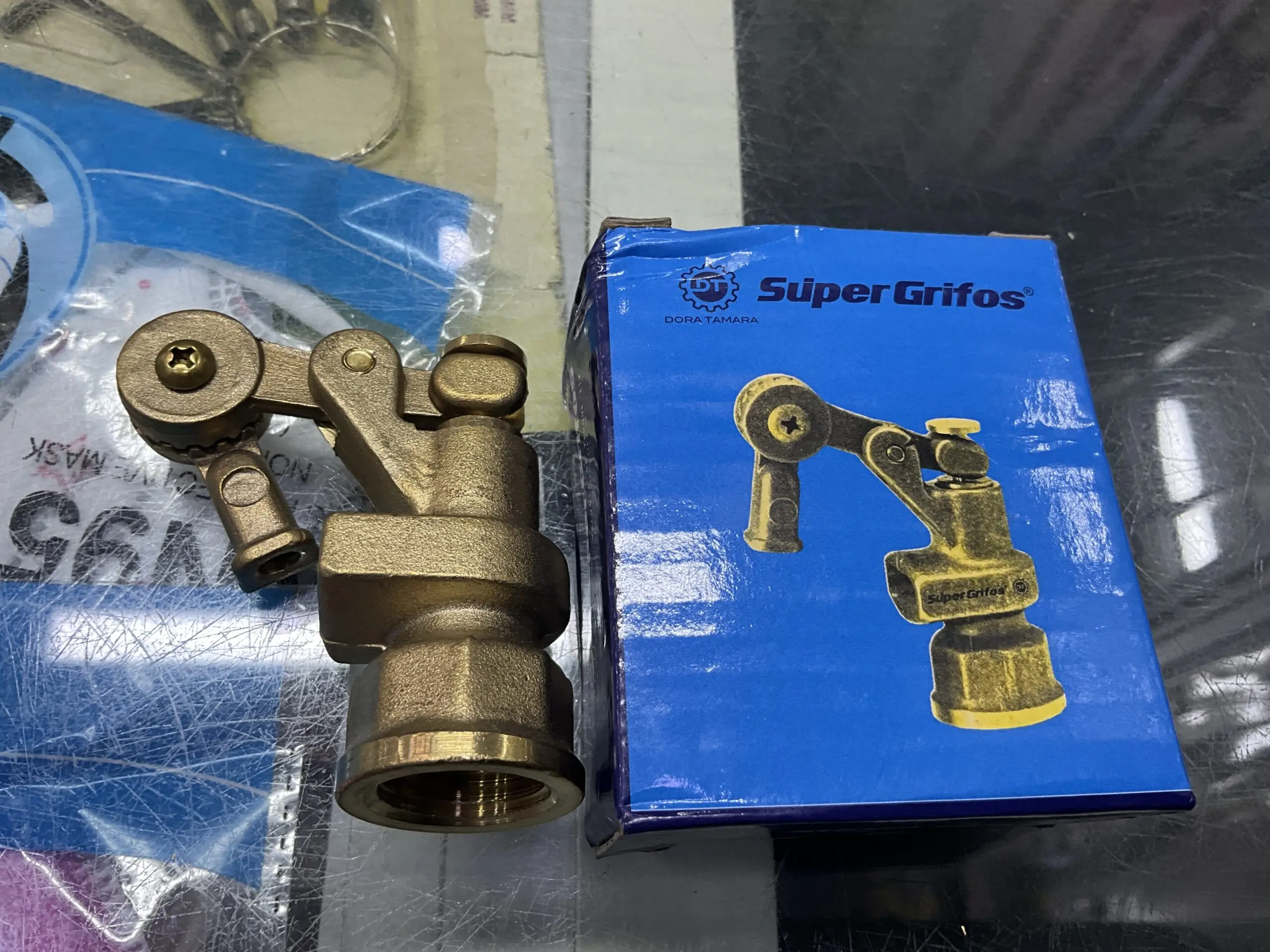 Válvula de tanque en bronce con brazo articulado junto a su caja azul de Super Grifos, resaltando su acabado metálico dorado y diseño mecánico.