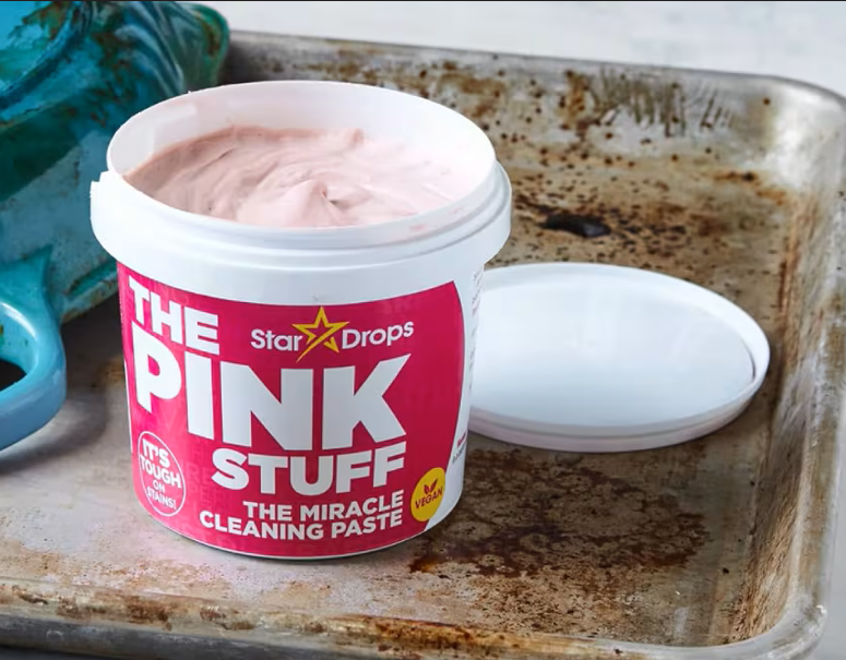Tarrina abierta de pasta limpiadora multiuso rosa con tapa blanca sobre bandeja metálica sucia y utensilio azul al fondo.