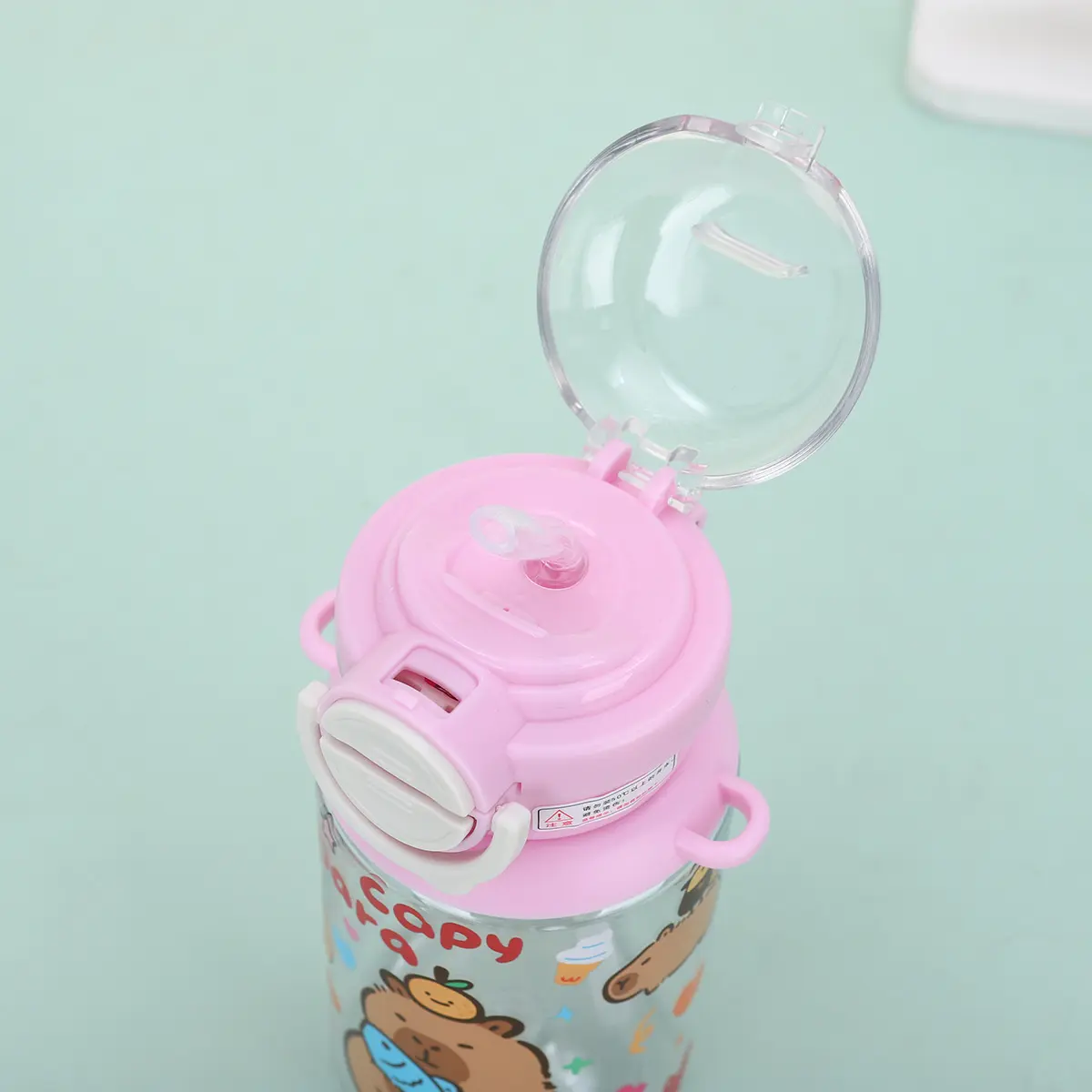 Botilito infantil rosa con tapa transparente abierta, pajilla de silicona y diseño decorativo de capibara para uso escolar.