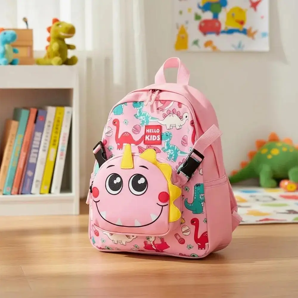 Morral escolar infantil rosa con estampado de dinosaurios y bolsillo frontal con cara de dinosaurio sonriente, en una habitación de niños.