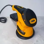 Lijadora eléctrica 3 en 1 rotoorbital amarilla y negra con control de velocidad, bolsa de polvo y cable, vista lateral sobre superficie gris.