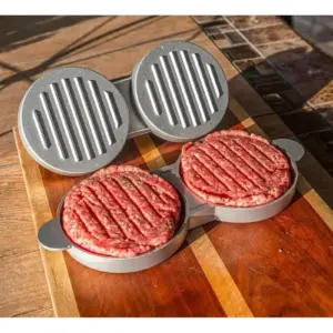 Prensa doble de hamburguesas de aluminio gris con tapas estriadas, mostrando dos carnes crudas sobre una tabla de madera bajo la luz del sol.