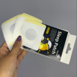 Mano sosteniendo caja de Sleep Patch con diez parches blancos cuadrados con centro circular gris y diseño de luna durmiendo