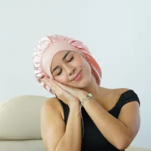Gorro de satín rosado pastel con diseño fruncido, usado por mujer descansando para proteger el cabello rizado.