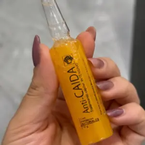 Ampolleta de tratamiento capilar intensivo Anti-Caída de 12 g con líquido naranja, sostenida por una mano femenina sobre fondo neutro.